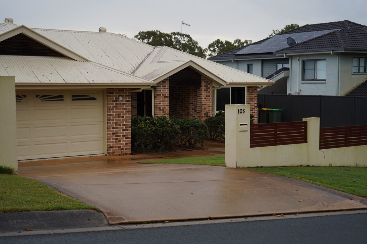 Wet plain concrete driveway with patchy tan staining sweeping past a brick home's cream double garage in Gumdale, Brisbane